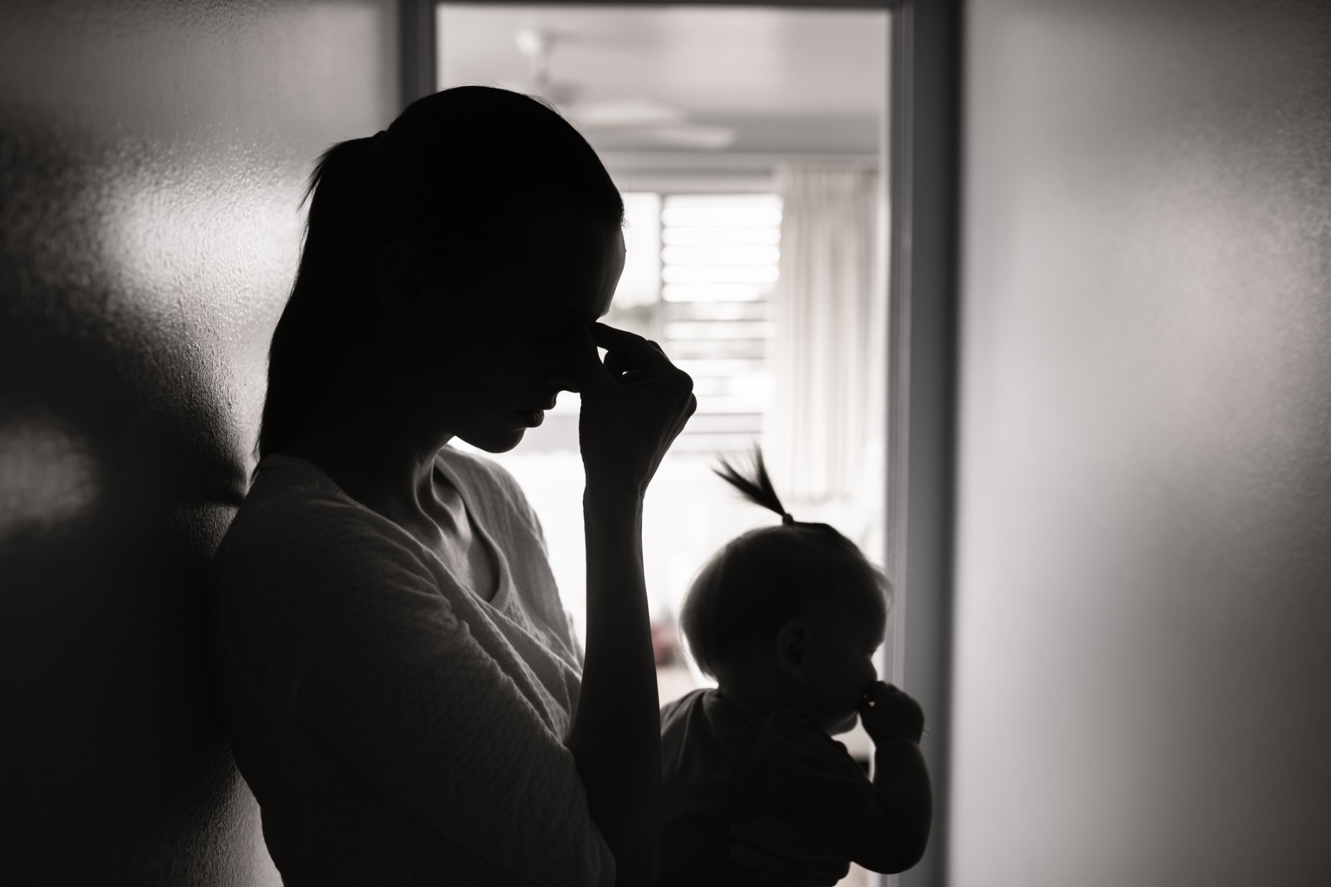 An overwhelmed and worried woman, leaning against a wall inside her home and holding a baby. 