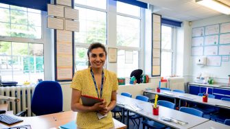 A medium closeup of a confident female teacher stood at the front of her classroom waiting for her pupils to arrive. 