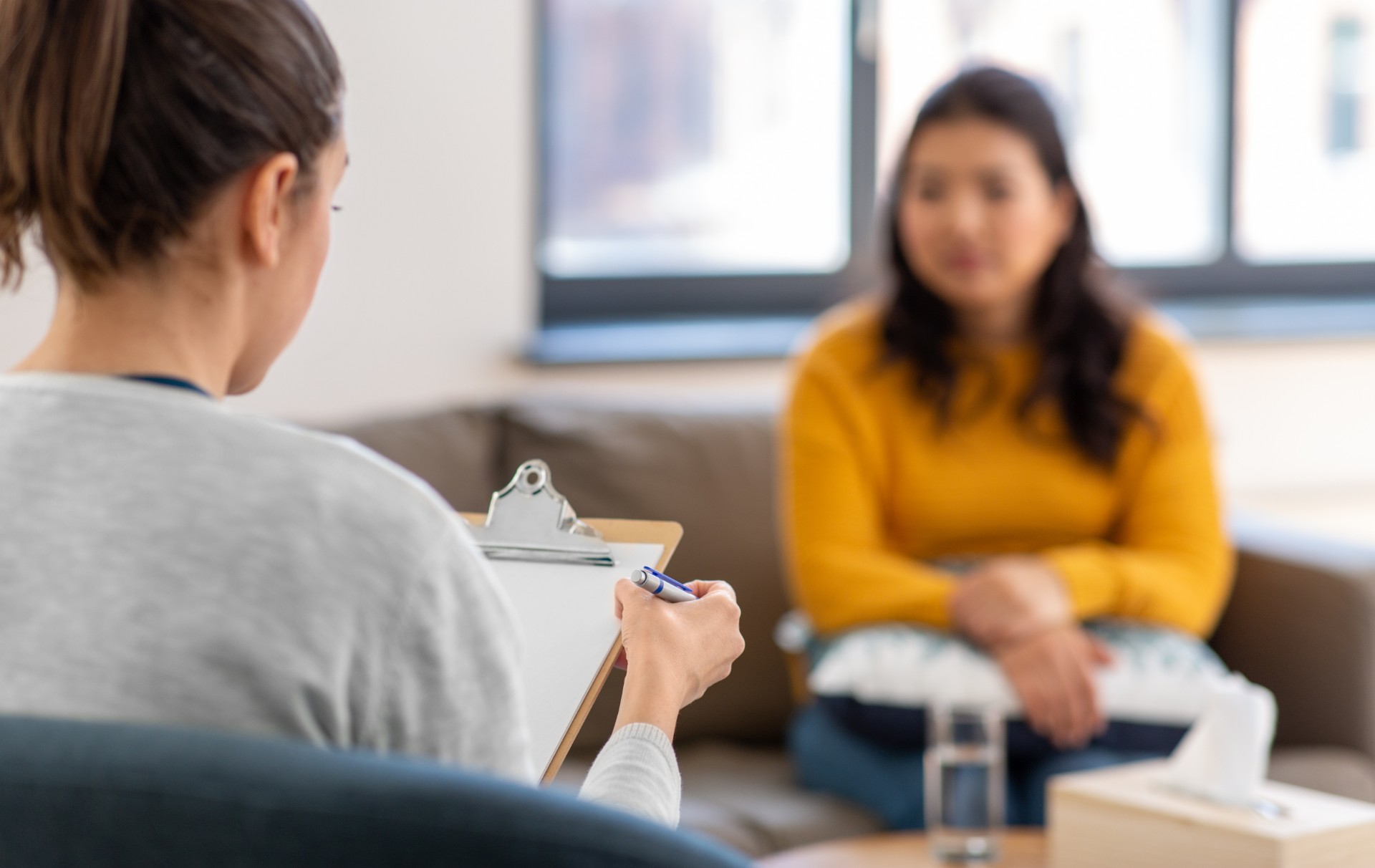 A patient sits on the couch opposite a therapist who is holding a clipboard.