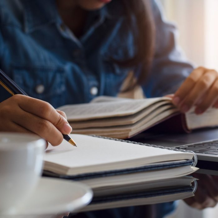 Casual young female student handwriting information on diary notebook while reading book and work on laptop computer with white cup of coffee on the table at home.