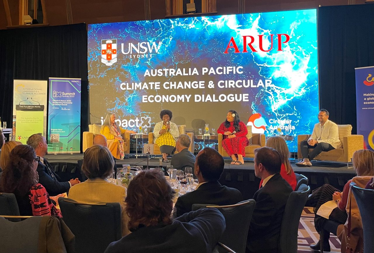 A panel of four people engage with each other on the stage, and with the audience in the foreground. There are three women and one man, in that order from left to right. They are people of colour from Australia, Papua New Guinea, Samoa, and Fiji. They are smiling and wear colourful clothing. The title of the conference is on the screen behind the panel members reads “Australia Pacific Climate Change & Circular Economy Dialogue”.