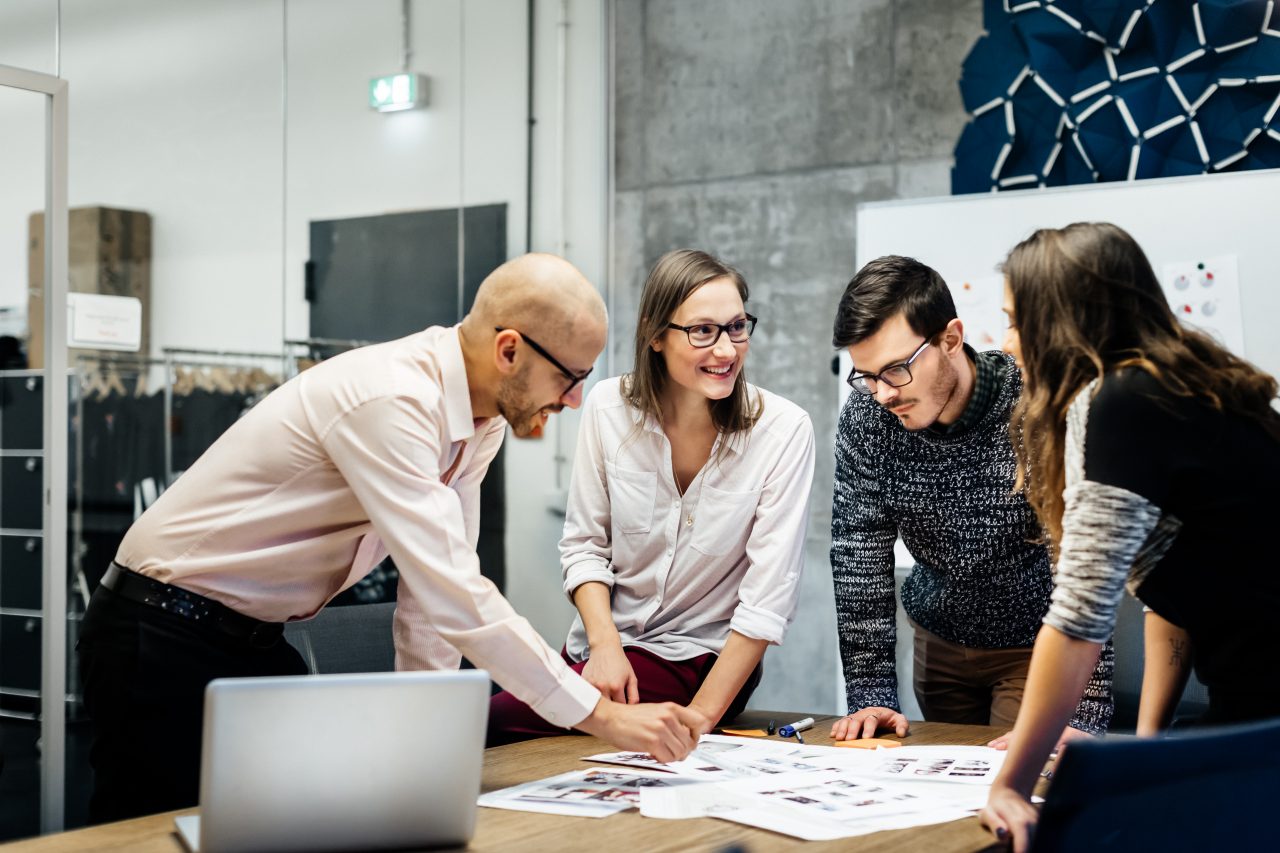 A team of four business people is standing in front a desk in a bright office room. They ambitiously point at documents while smiling and looking at the charts and notes.