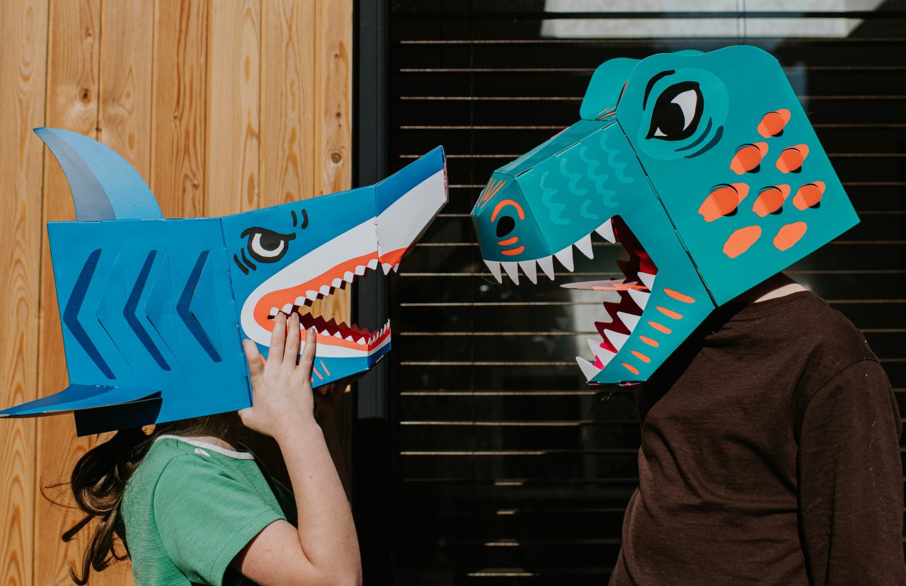 Two kids standing face-to-face wearing homemade, bright masks of a shark and a dinosaur baring their teeth.