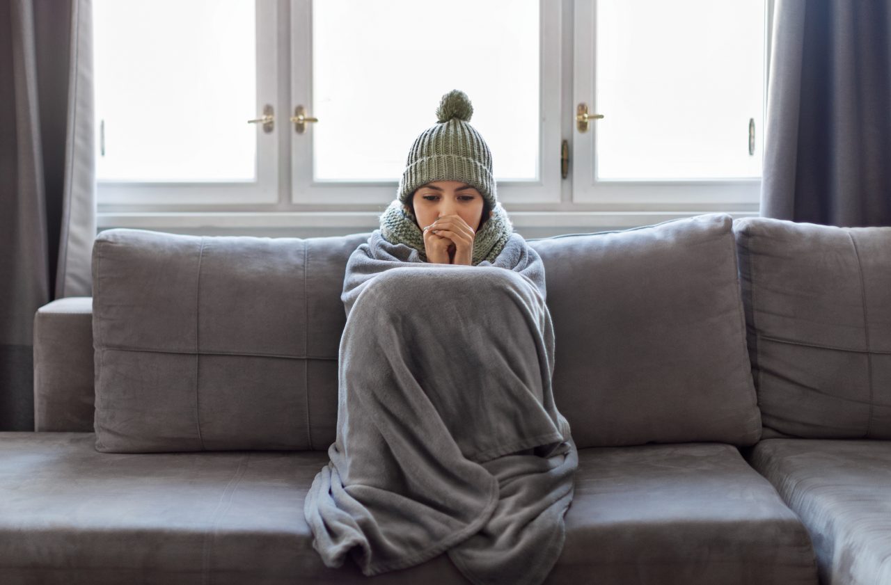 Cold Home. Young Woman Covered With Blanket Freezing On Couch In Living Room, Millennial Female Wearing Knitted Scarf And Hat Indoors Warming Hands With Her Breath, Suffering Heating Problems