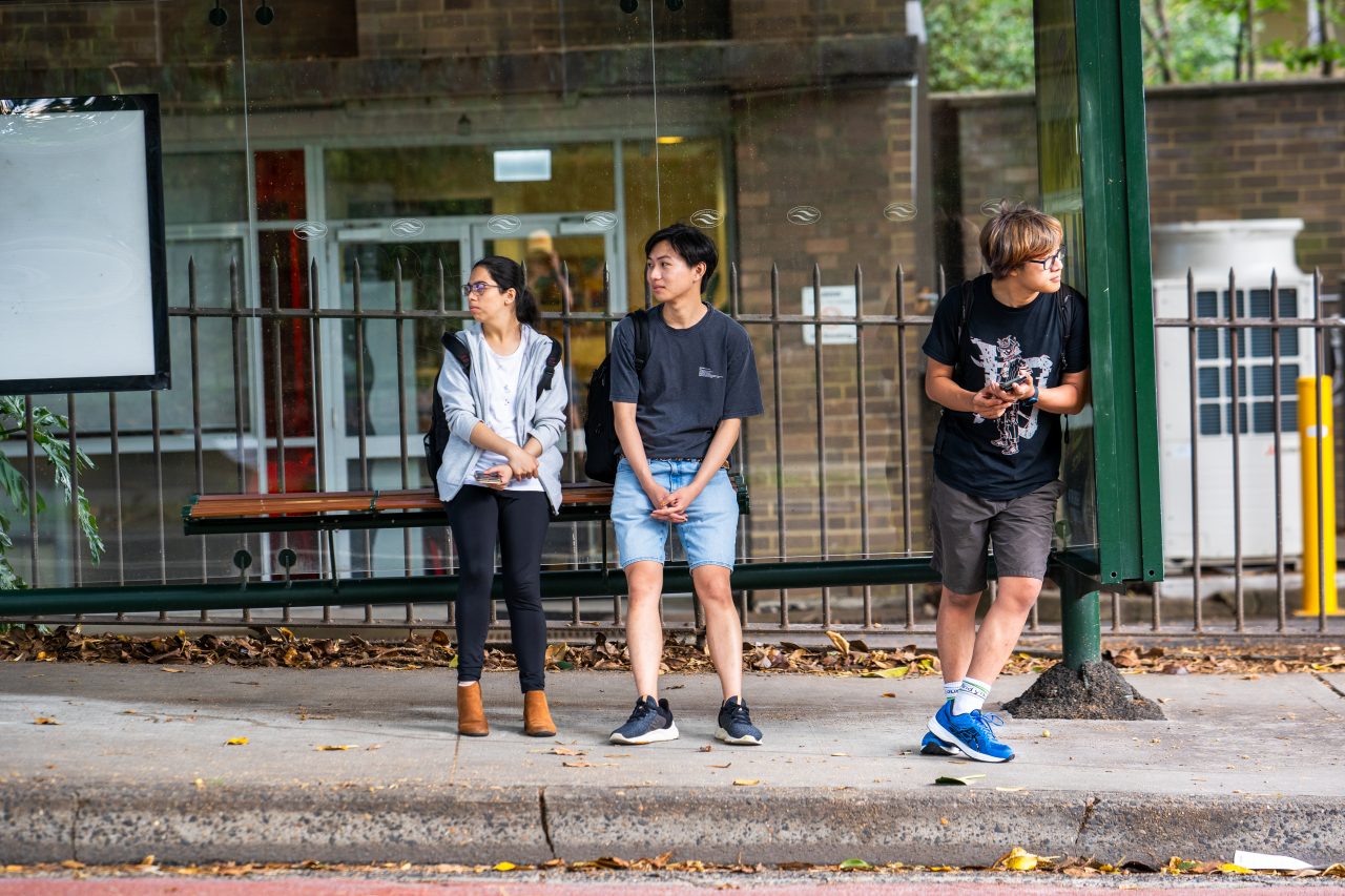 Students sitting at a bus stop on High Street outside Kensington Campus