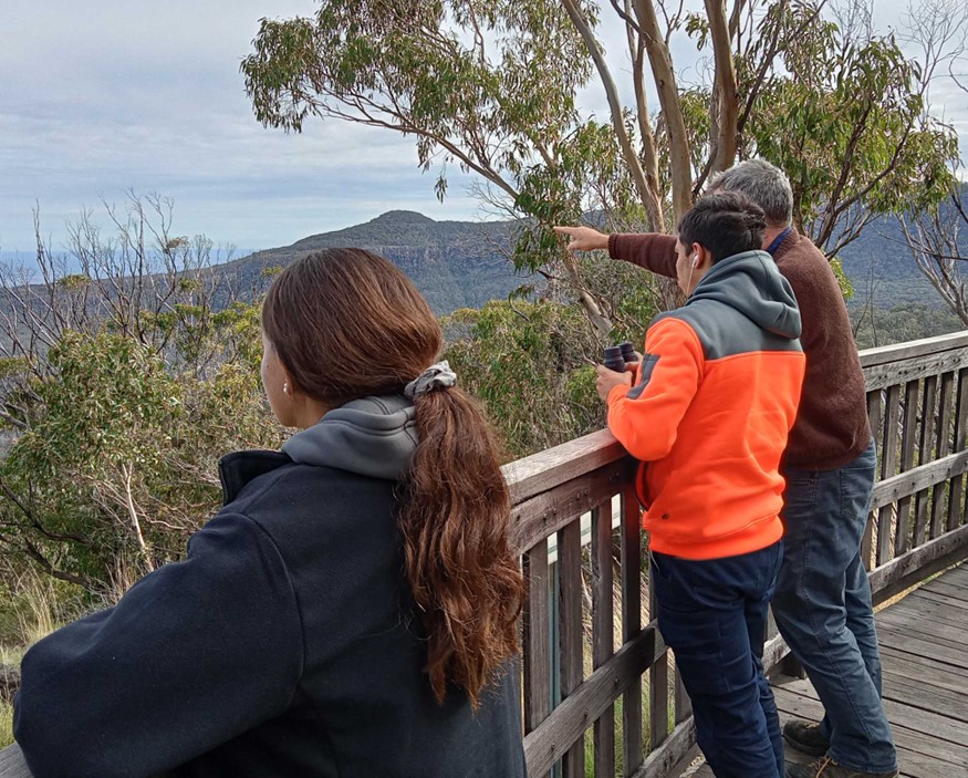 Three people with their backs to the camera are looking out on Australian bush on a wooden platform